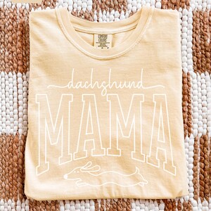 May include: A light yellow t-shirt with the words "dachshund MAMA" in white script and block letters. A white outline of a dachshund dog is below the word "MAMA". The shirt is made of a soft material.