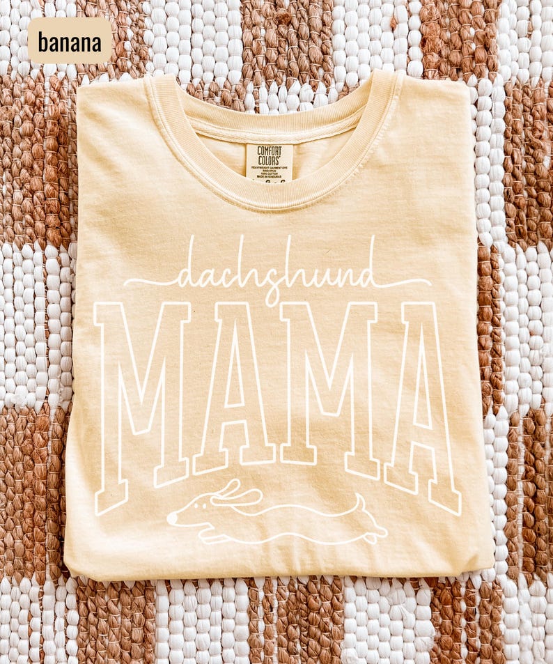 May include: A light yellow t-shirt with the words "dachshund MAMA" in white script and block letters. A white outline of a dachshund dog is below the word "MAMA". The shirt is made of a soft material.