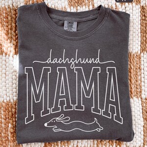 May include: Pepper-colored Comfort Colors t-shirt with the words "dachshund MAMA" in white, along with a line drawing of a dachshund. The shirt has a crew neck and short sleeves.
