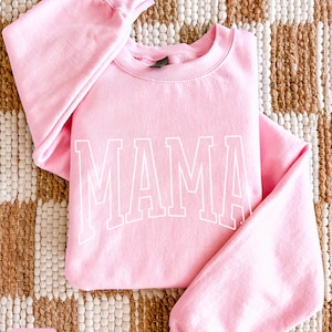 May include: A pale pink sweatshirt featuring the word "MAMA" in white, block lettering. The garment has a crew neck and long sleeves. The background is a woven rug in neutral tones.