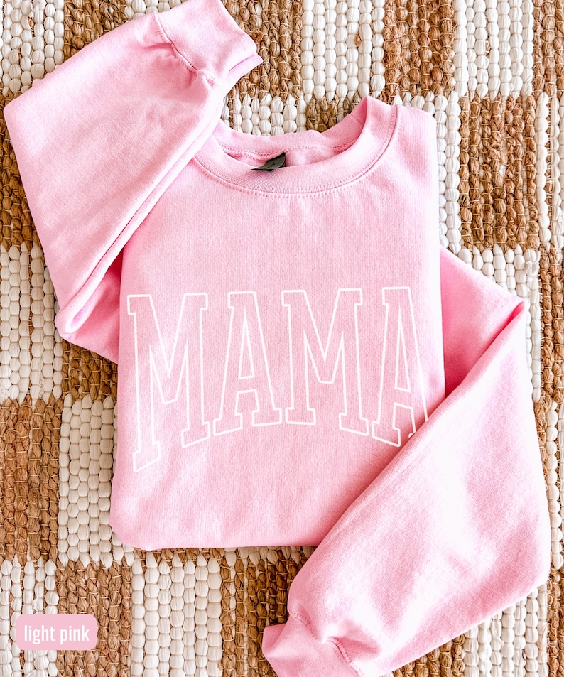 May include: A pale pink sweatshirt featuring the word "MAMA" in white, block lettering. The garment has a crew neck and long sleeves. The background is a woven rug in neutral tones.