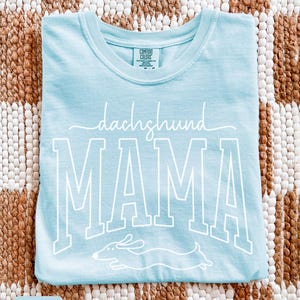 May include: Light blue t-shirt with the words "dachshund MAMA" in white script and block letters. A white dachshund dog graphic is below the word "MAMA". The shirt is made of a soft material.