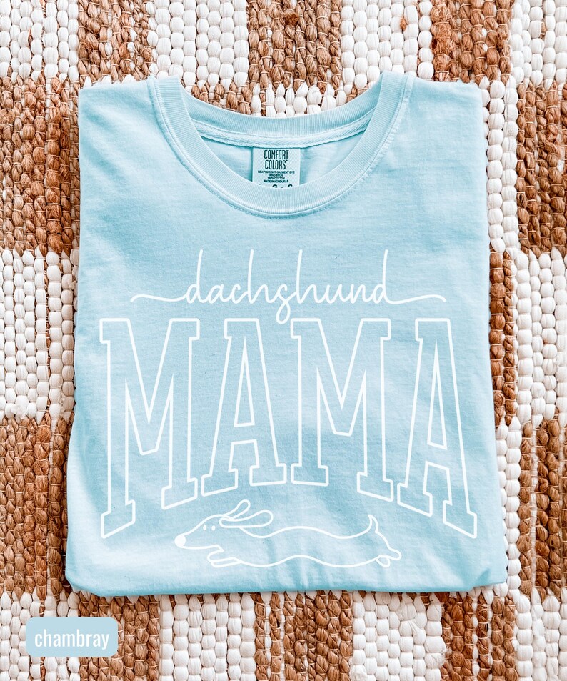 May include: Light blue t-shirt with the words "dachshund MAMA" in white script and block letters. A white dachshund dog graphic is below the word "MAMA". The shirt is made of a soft material.