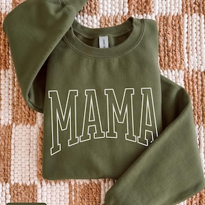 May include: A military green sweatshirt featuring the word "MAMA" in white block lettering. The crew neck sweatshirt is made of a soft material. The long-sleeved top is a casual style.