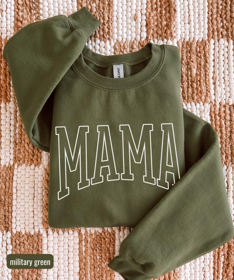 May include: A military green sweatshirt featuring the word "MAMA" in white block lettering. The crew neck sweatshirt is made of a soft material. The long-sleeved top is a casual style.