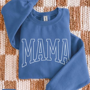 May include: Indigo blue sweatshirt featuring the word "MAMA" in white block lettering. The crew neck sweatshirt has long sleeves. The text "indigo blue" is in a small box in the lower left corner.