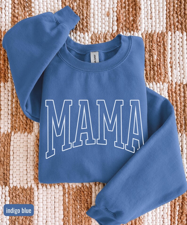 May include: Indigo blue sweatshirt featuring the word "MAMA" in white block lettering. The crew neck sweatshirt has long sleeves. The text "indigo blue" is in a small box in the lower left corner.