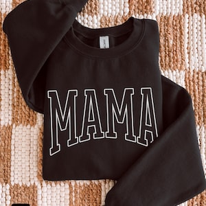 May include: Black sweatshirt with the word "MAMA" in white, block letters. The sweatshirt is folded on a textured, neutral-toned surface. The word "black" is in a small box in the lower left corner.