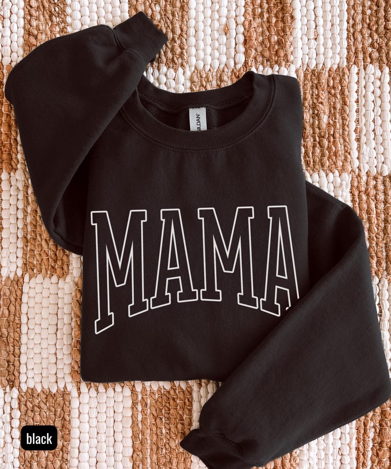 May include: Black sweatshirt with the word "MAMA" in white, block letters. The sweatshirt is folded on a textured, neutral-toned surface. The word "black" is in a small box in the lower left corner.