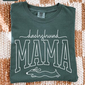 May include: Blue spruce Comfort Colors t-shirt with the words "dachshund MAMA" in white block letters and a dachshund illustration. The shirt is a crew neck and has a tag on the back of the neck.
