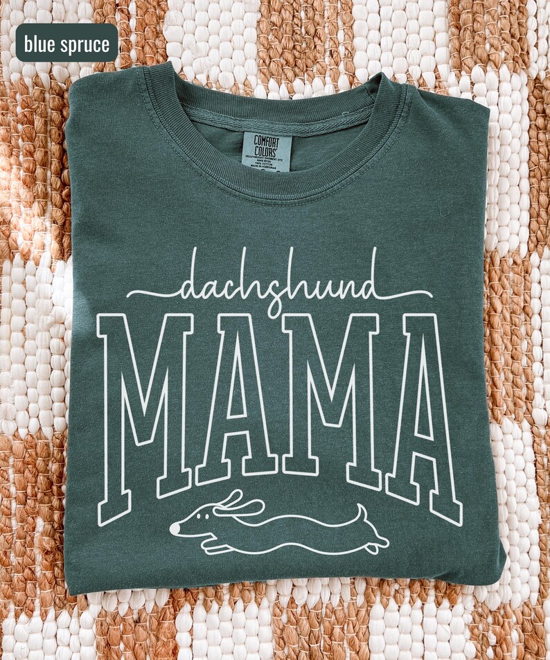 May include: Blue spruce Comfort Colors t-shirt with the words "dachshund MAMA" in white block letters and a dachshund illustration. The shirt is a crew neck and has a tag on the back of the neck.