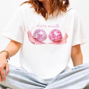 May include: White t-shirt with a playful illustration of two pink snails, each with a disco ball shell. The words "disco snails" are written above the snails. A casual, everyday wear item.
