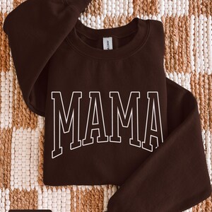 May include: A dark chocolate-coloured sweatshirt featuring the word "MAMA" in white, outlined block letters. The garment is folded on a textured, neutral-toned surface. The words "dark chocolate" are in a small white box at the bottom.