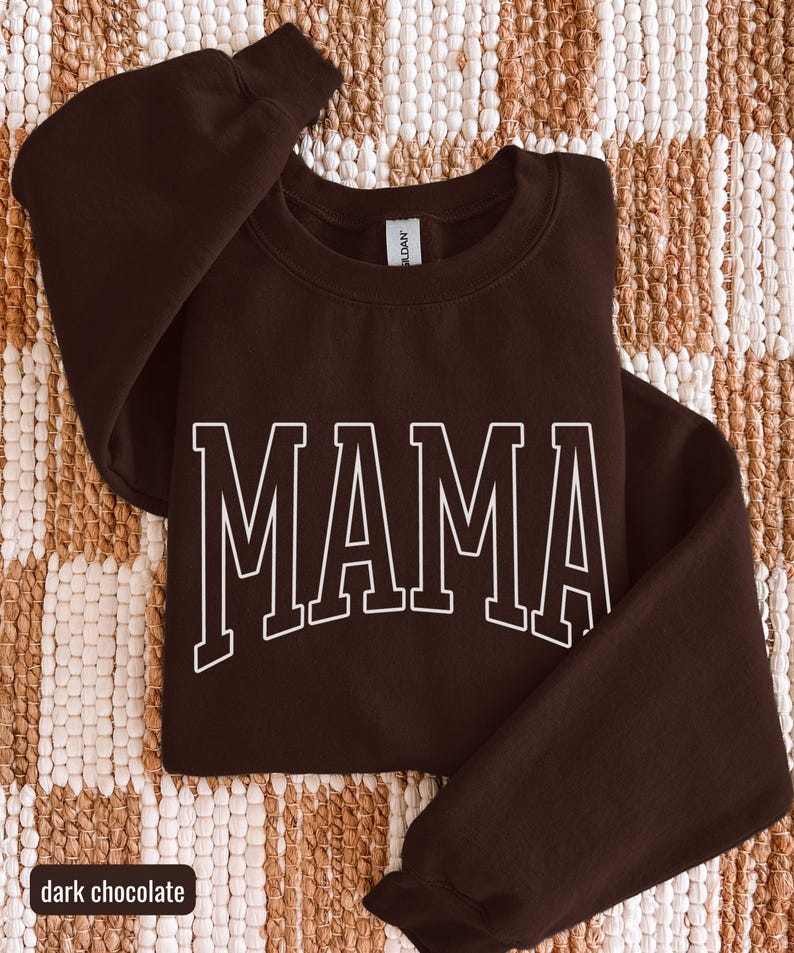 May include: A dark chocolate-coloured sweatshirt featuring the word "MAMA" in white, outlined block letters. The garment is folded on a textured, neutral-toned surface. The words "dark chocolate" are in a small white box at the bottom.
