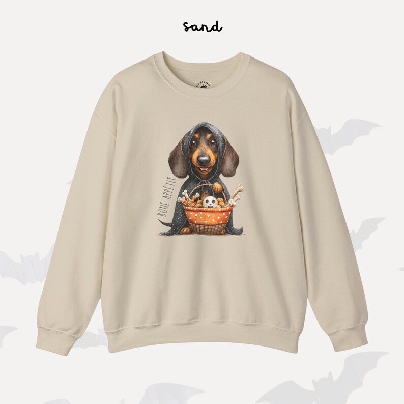 May include: A sand-coloured sweatshirt with a dachshund wearing a hooded cloak and holding a basket of bones and a small ghost. The words "BONE APP&Eacute;TIT" are printed on the left side of the dog. The word "sand" is printed at the top.