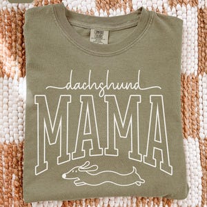 May include: Khaki-colored t-shirt with the words "dachshund MAMA" in white lettering and a dachshund illustration. The shirt is made of a soft material and has a crew neck.