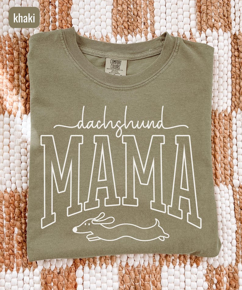 May include: Khaki-colored t-shirt with the words "dachshund MAMA" in white lettering and a dachshund illustration. The shirt is made of a soft material and has a crew neck.