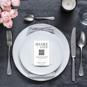 Op de afbeelding: Een witte kaart met zwarte tekst die "SHARE the LOVE" zegt en een QR-code heeft. De tekst onder de QR-code zegt "SCAN OUR QR CODE TO UPLOAD YOUR BEST PICTURES & VIDEOS TO OUR DIGITAL ALBUM with love EMILY & NICK 11.11.20XX"