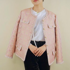 May include: A light pink jacket with a gold chain pattern, round collar, and gold buttons. The jacket has two front pockets and a fringed trim. The model is wearing a white shirt and black velvet pants.