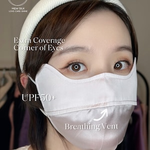 May include: A light pink silk face mask with a breathing vent and UPF 50+ protection. The mask has extra coverage around the eyes and full coverage of the nose and mouth. The text "Extra Coverage Corner of Eyes", "UPF 50+", "Breathing Vent", and "Full Coverage" is written on the mask.