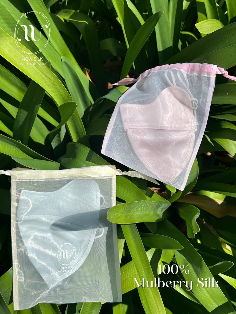 May include: Two silk face masks in separate white mesh bags. The mask on the left is light blue and the mask on the right is light pink. Both bags have a white logo with the text "M&W SILK LOVE CARE SHINE". The text "100% Mulberry Silk" is written below the bags.