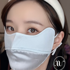 May include: A light blue silk face mask with a white headband. The mask has a unique design with a curved top and a lower section that covers the chin. The mask is made of two layers of silk, with the top layer being a solid color and the bottom layer being a satin finish. The headband is made of a soft, comfortable material and is designed to be worn comfortably on the head. The mask is designed to be worn over the nose and mouth to help protect against the spread of germs.