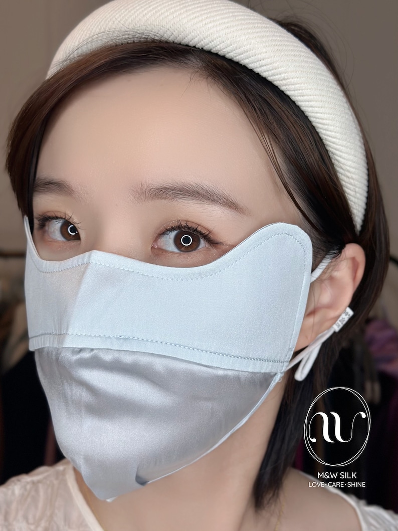 May include: A light blue silk face mask with a white headband. The mask has a unique design with a curved top and a lower section that covers the chin. The mask is made of two layers of silk, with the top layer being a solid color and the bottom layer being a satin finish. The headband is made of a soft, comfortable material and is designed to be worn comfortably on the head. The mask is designed to be worn over the nose and mouth to help protect against the spread of germs.