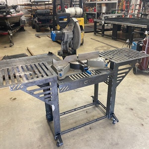 May include: A black metal miter saw stand with two side tables and wheels. The stand has a metal grate on the top and a metal frame with a cut-out design. The stand is in a workshop setting.