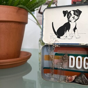 Op de afbeelding: Een vintage blikken doos met een zwart-witte illustratie van een puppy op de deksel. De doos is open en de binnenkant is versierd met kleurrijk washi tape en een klein glazen flesje met gedroogde bloemen. Het woord "DOG" is in witte letters op de bodem van de doos geschreven.