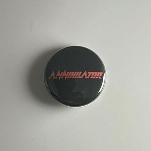May include: A round, black button with the word "ANNIHILATOR" in red, stylized text. The button has a glossy finish and is centered on a white background. The text appears to be a band or brand name.