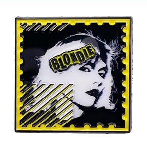 May include: Square pin featuring a black and white image of a woman with the word "BLONDIE" in a banner. The pin has a yellow border with a postage stamp design and yellow diagonal stripes.