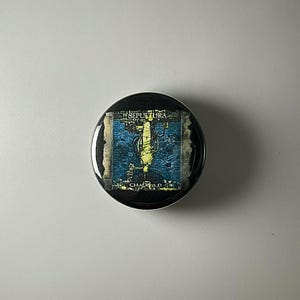 May include: A circular pin-back button featuring the Sepultura Chaos A.D. album cover art. The design displays a central yellow figure against a dark blue and black background with the text Sepultura at the top and Chaos A.D. at the bottom.