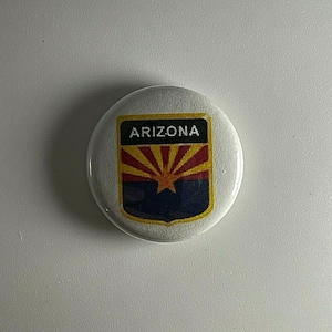 Può includere: Un bottone rotondo bianco con la scritta "ARIZONA" in alto. Il bottone presenta il design della bandiera dell'Arizona, con un sole giallo su sfondo blu e un bordo dorato.