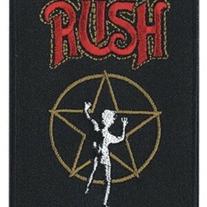 May include: A rectangular black embroidered patch featuring the red text RUSH at the top. Below the text is a gold-colored star outline with a white silhouette of a person standing inside the star with arms raised in a pose.