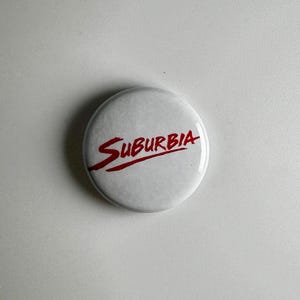 Peut inclure: Un bouton blanc avec le mot "Suburbia" en script rouge. Le bouton est rond et a une finition brillante. Le texte est en cursive.