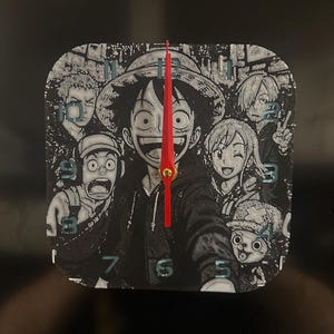 May include: Square wall clock featuring a black and white manga-style illustration of several anime characters. The clock face has a red second hand and blue numerals. The characters are smiling and looking at the viewer.