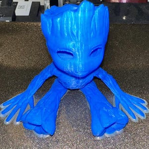 May include: A blue 3D printed figurine of a character from the Guardians of the Galaxy movie franchise. The character is known as Groot and is depicted in a sitting position with outstretched arms and hands.