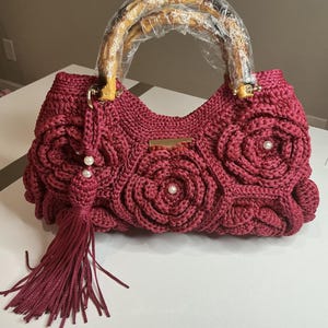 May include: A burgundy crochet handbag with a bamboo handle. The bag features a floral design with pearl accents and a matching tassel. The handle is wrapped in clear plastic. The bag is sitting on a white surface.