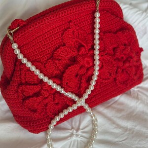 May include: A vibrant red crochet handbag with a floral design. The bag features a pearl beaded strap and gold-colored hardware. The bag is on a white background.