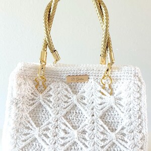 May include: A white crocheted handbag featuring a diamond-patterned texture, two braided gold-tone handles, and a rectangular gold-tone metal plaque centered on the front with the text D' Gertru engraved in a script font.