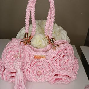 May include: A light pink crocheted handbag with a floral rose design. The bag features a braided handle, gold-colored hardware, and a tassel with pearl accents. The bag is sitting on a white surface.
