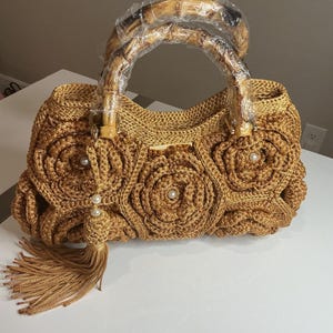 May include: A gold-colored crochet handbag with a bamboo handle. The bag features a floral design with pearl accents and a tassel. The handle is wrapped in clear plastic. The bag is sitting on a white surface.