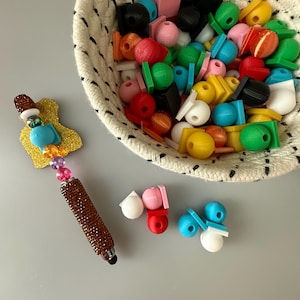 May include: A selection of brightly coloured beads and shapes, including spheres, cubes, and cylinders, are displayed in a woven basket and scattered on a grey surface. A decorative pen with a brown beaded barrel and a gold glitter accent is also present.
