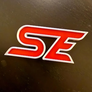 May include: Red and silver "SE" logo with a stylized font. The letters are raised and have a 3D effect.