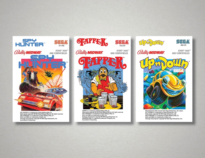 Pu&ograve; includere: Tre scatole di videogiochi vintage per console Atari 2600. Le scatole presentano grafica e testo colorati, tra cui "Spy Hunter", "Tapper" e "Up 'n Down". La scatola di "Spy Hunter" mostra un'auto rossa e delle figure. La scatola di "Tapper" ha un barista. La scatola di "Up 'n Down" ha un'auto gialla.
