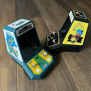 以下が含まれることがあります： 2つのビンテージの携帯用ビデオゲーム機。左側の青と緑のゲーム機は、画面にゲーム「ギャラクシアン」と「コレコ」の文字が表示されています。右側の黒と黄色のゲーム機は、画面にゲーム「パックマン」と「ミッドウェイのパックマン」の文字が表示されています。