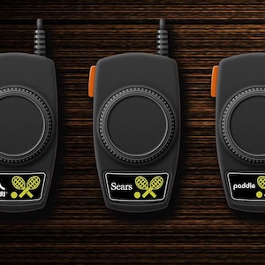 May include: Three black Atari paddle controllers with orange accents and a circular design. Each controller has a different label: Atari, Sears, and paddle. The controllers have a tennis ball and racket graphic.