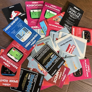 May include: A collection of vintage Activision video game cartridges, featuring titles like "Kaboom!", "Fishing Derby", and "Kung-Fu Master". The cards display colorful graphics and game titles, with the Activision logo prominently featured.