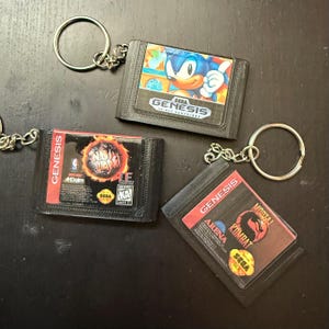 Op de afbeelding: Drie zwarte Sega Genesis-spelcartridge-sleutelhangers. De eerste sleutelhanger heeft een blauw Sonic the Hedgehog-personage. De tweede sleutelhanger heeft een rood, geel en zwart NBA Jam-logo. De derde sleutelhanger heeft een rood Mortal Kombat Arena-logo met een zwarte draak.