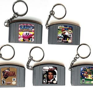 May include: Five Nintendo 64 game cartridge keychains. The cartridges feature the games Kirby's Crystal Shards, Mario Kart 64, The Legend of Zelda: Ocarina of Time, GoldenEye 007, and Super Mario 64.